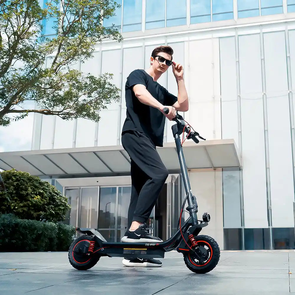 Un homme portant des lunettes de soleil debout sur une trottinette électrique T10 Pro noire avec des accents rouges devant un immeuble de bureaux moderne.