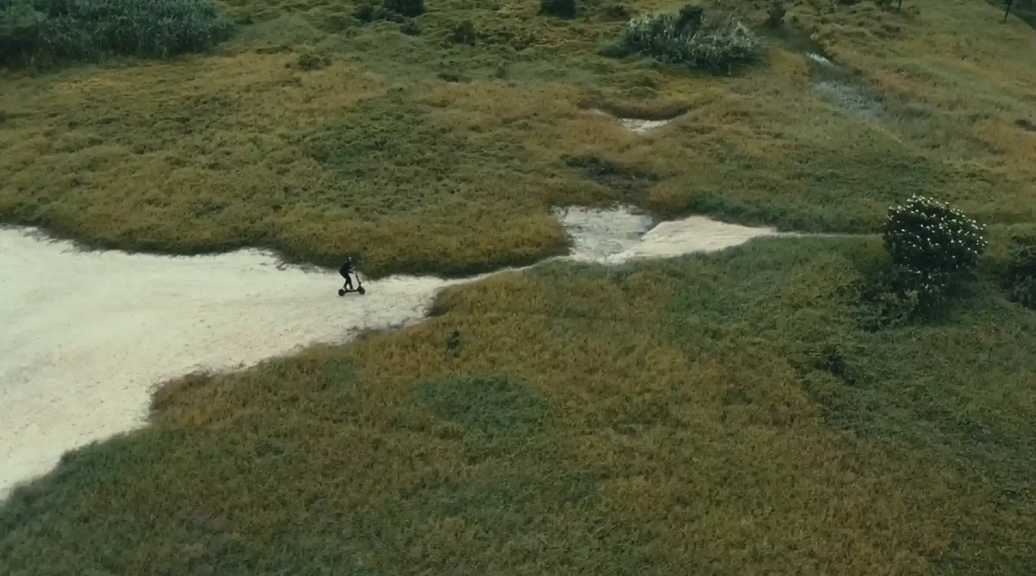 Vue aérienne d'une personne pilotant une trottinette électrique tout-terrain sur un chemin sablonneux à travers une vaste étendue de végétation sauvage.